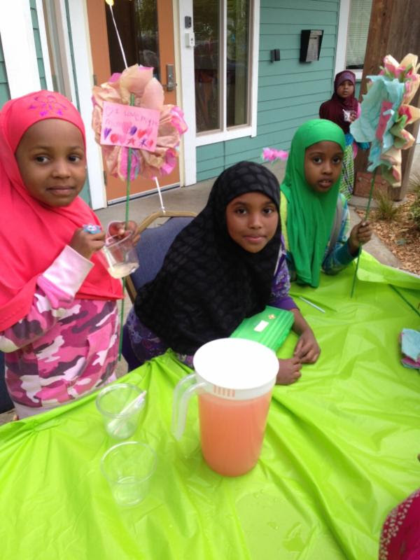 Children smiling at the camera while sitting at a table with a pitcher of lemonade