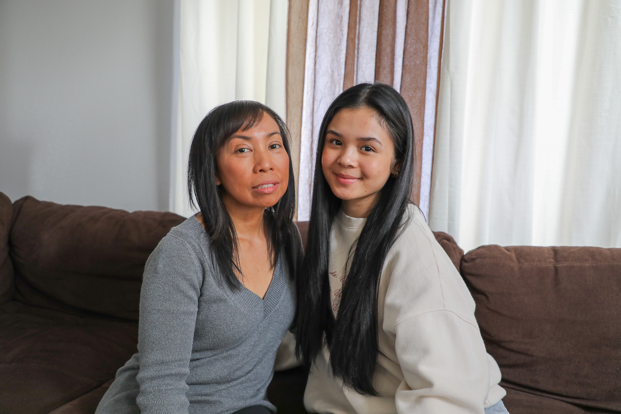 photo of foster parent Daisy (left) and her foster child, Jas (right), seated on a brown couch with a window covered by white and maroon curtains in the background.