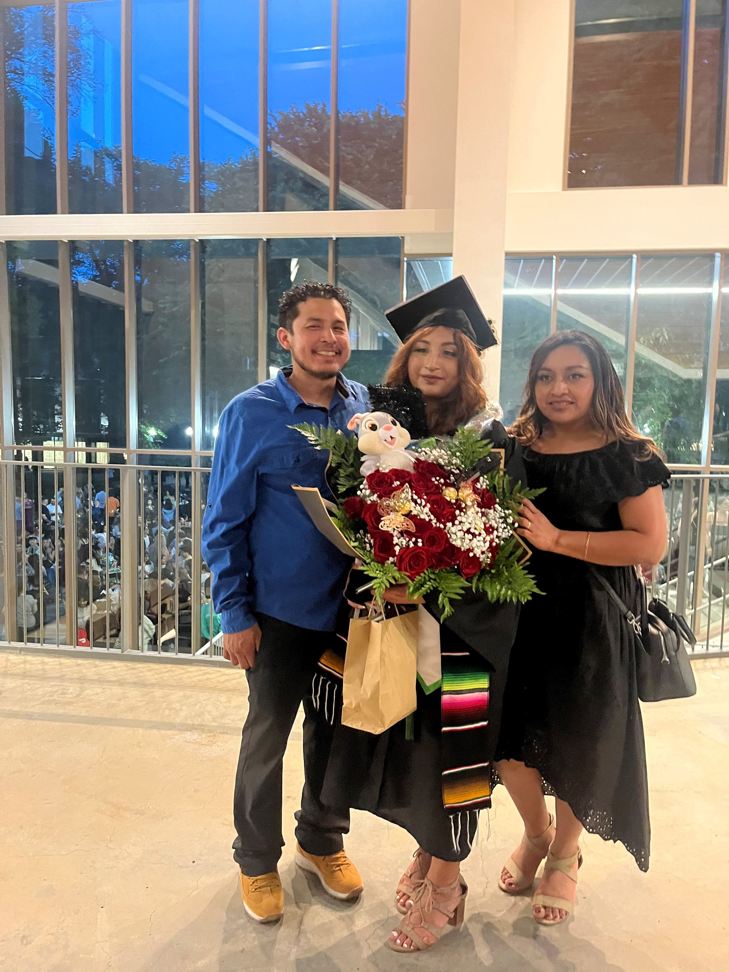 Mayel Interian-Ku (center) stands in her graduation gown at the Parkrose High School graduation ceremony in June 2023, with her father on her right side and her mother on her left side. Mayel holds a giant bouquet of flowers and a brown paper bag. Mayel, her mother, and her younger siblings all participated in the Morrison’s Listos Para Aprender early learning program when they were young children. 