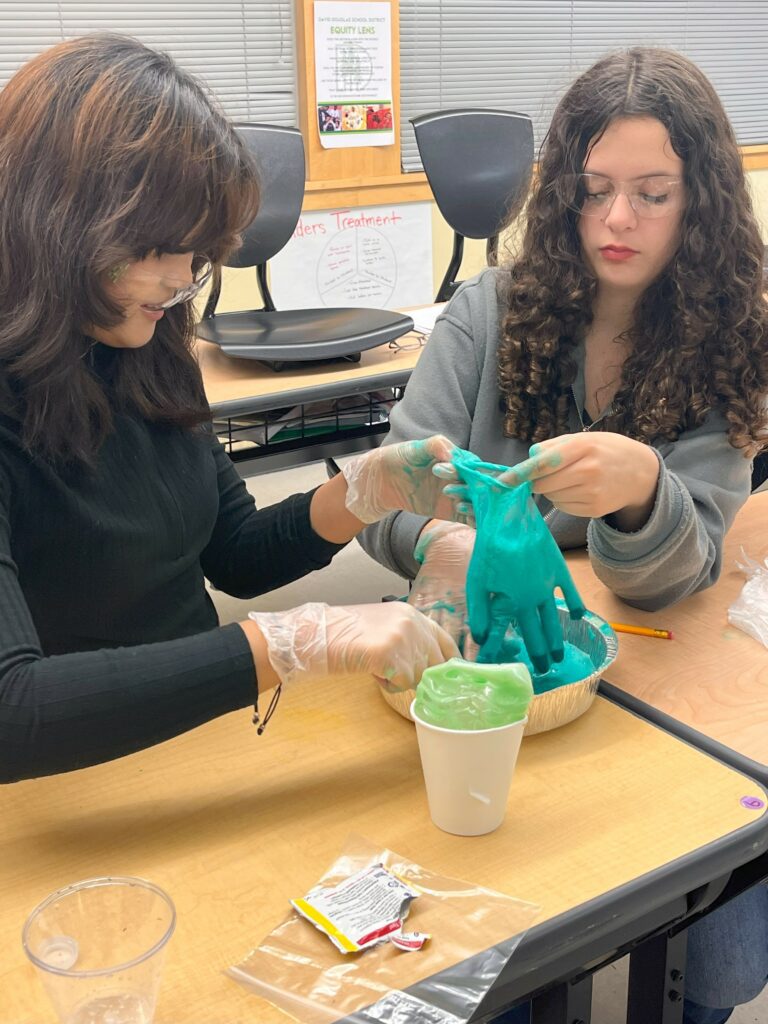 Thi Ri (left) in a black, long-sleeved shirt, with Shadae (right), in a gray long-sleeved shirt, sitting at desks next to each other at Ron Russell Middle School. Each student holds a latex glove with their left hand while using their right hand to fill it with an aquamarine substance in a round, tinfoil pan as part of a science experiment. A white paper cup with a light green item, a Ziploc bag and a torn packet sit in the foreground.