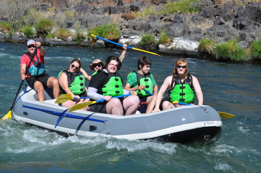 Five youths from New Avenues for Youth sit inside a gray raft on a river in Central Oregon. They are wearing green life vests and carrying blue and white oars. A guide in a red shirt and gray baseball cap sits at the back of the raft with an oar.