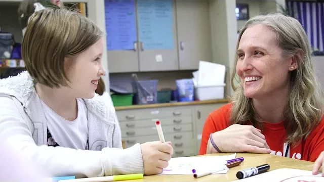 A mentor named Gretchen and a young girl smile at each other while coloring together during Girls Inc