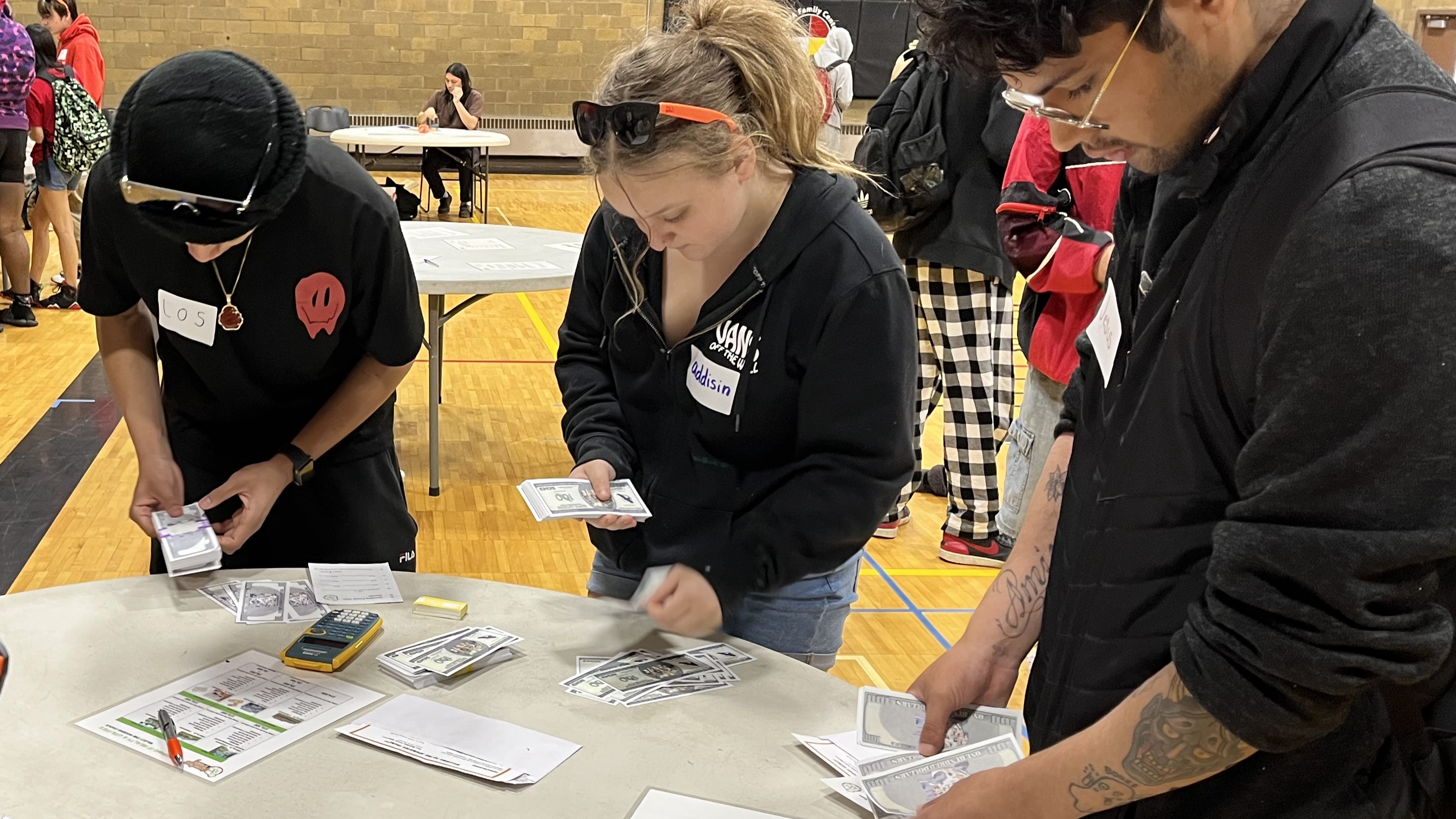 Photo of students in NAYA's college and career prep program seated and standing around a circular table looking at papers and spending pretend money during a budgeting exercise. Another half dozen people sit and stand around tables in the background of the gym.