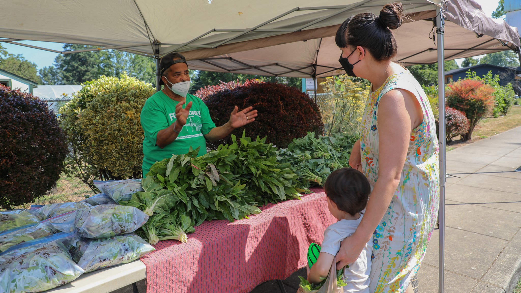 Photo of Our Village Gardens volunteer standing behind a table covered in vegetables they are distributing for free to people at an event in the New Columbia community in North Portland. The volunteer has hands extended in front of him while he explains one way to prepare a vegetable to a mother with a young child on the other side of the table.