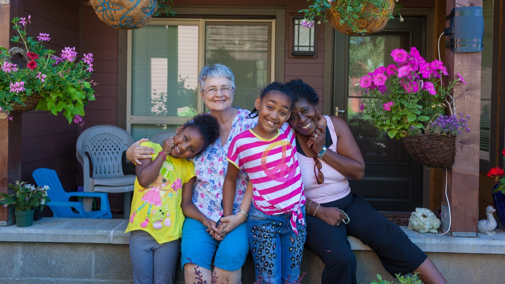 photo of two adults and two children sitting and standing in front of a front porch with flower pots on either side. photo courtesy of Bridge Meadows