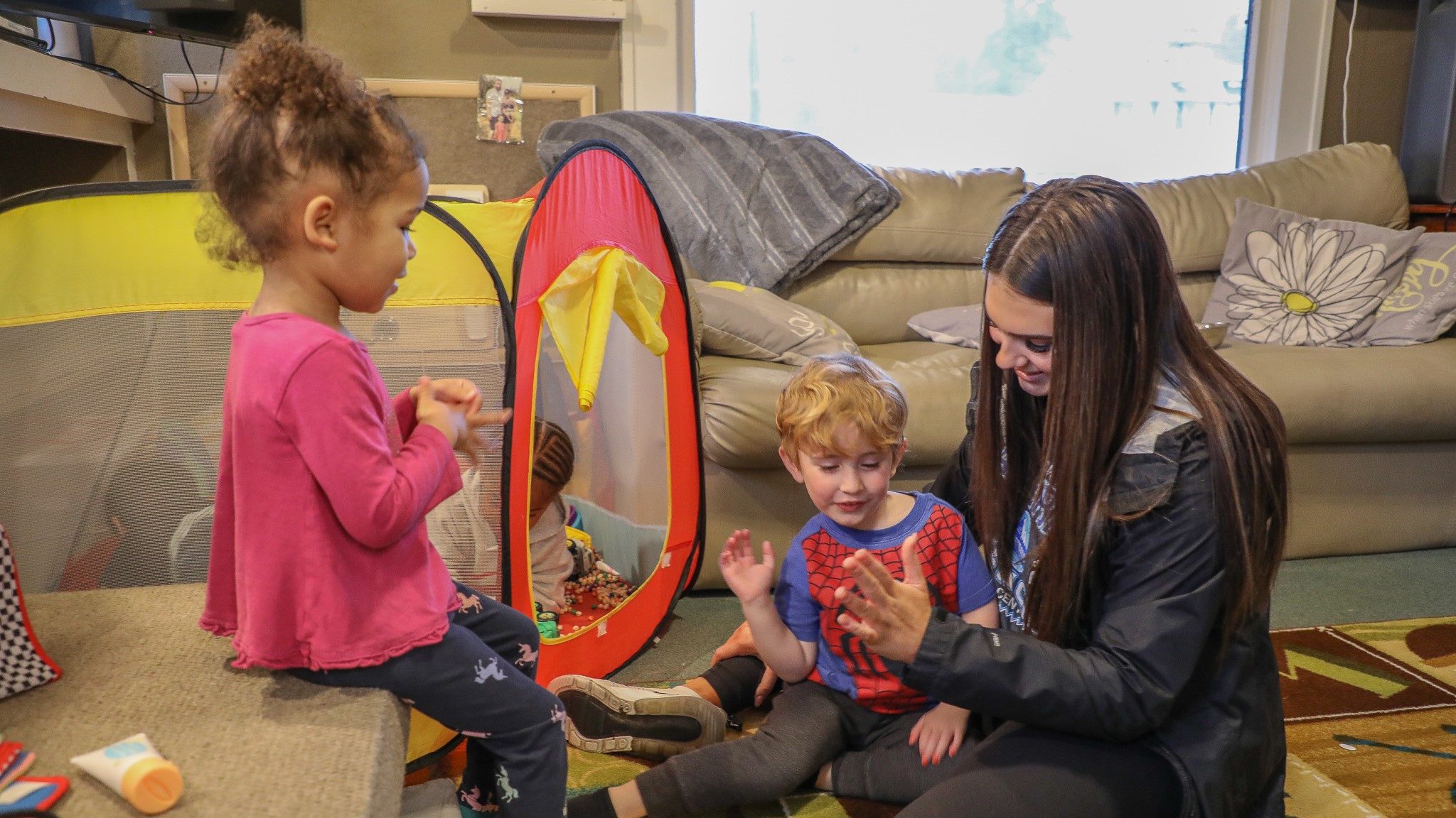 photo of a preschool staffer sitting on the floor and exchanging high fives with a child while another child sits on a couch in front of them