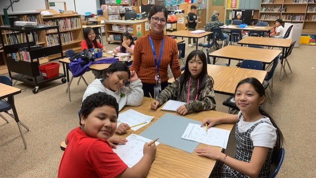 Photo of an IRCO youth advocate smiling at the camera while standing behind a square table with four students. Other students sit at square tables in the background, and shelves of books line the walls.