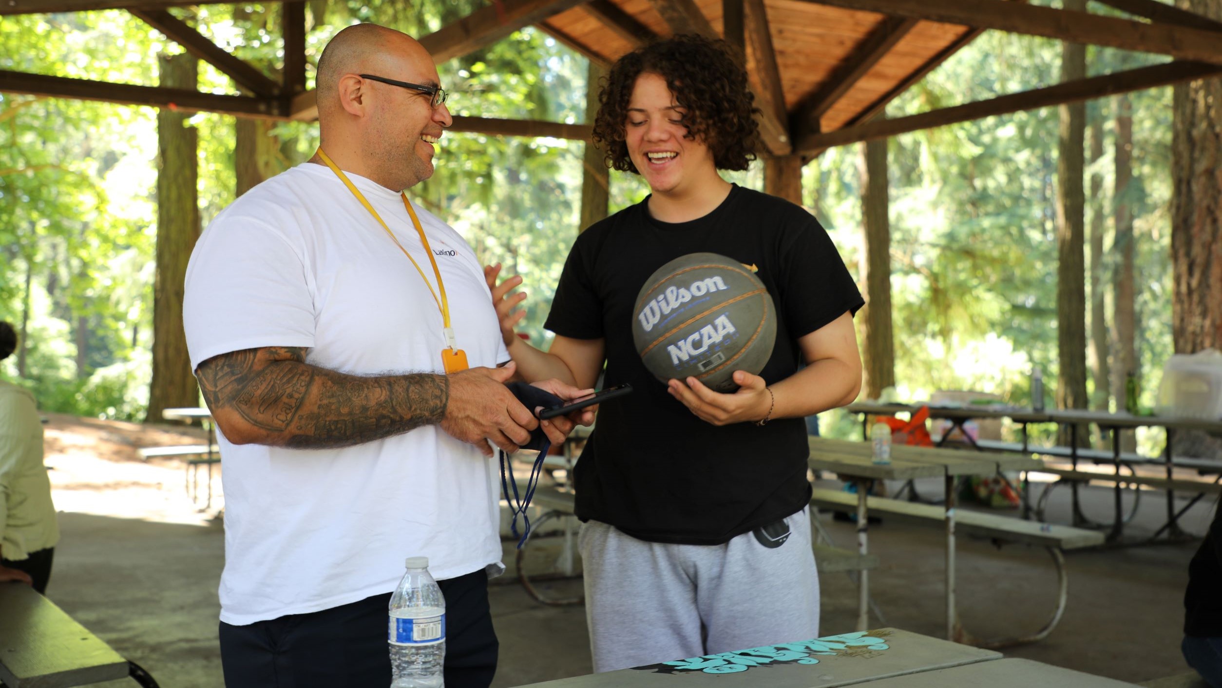 photo of Latino Network Padrinos mentoring program mentor and mentee at Mount Tabor Park. Both are standing near each other as they speak and smile, and mentee is holding a basketball.