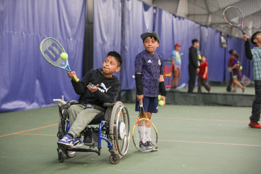 photo of James John Elementary School students using racquets to practice serves while on a court at Portland Tennis & Education. One student on the left uses a wheelchair while practicing their serve, while a friend stands by watching with a racquet and tennis balls. Other students and coaches stand in the background.