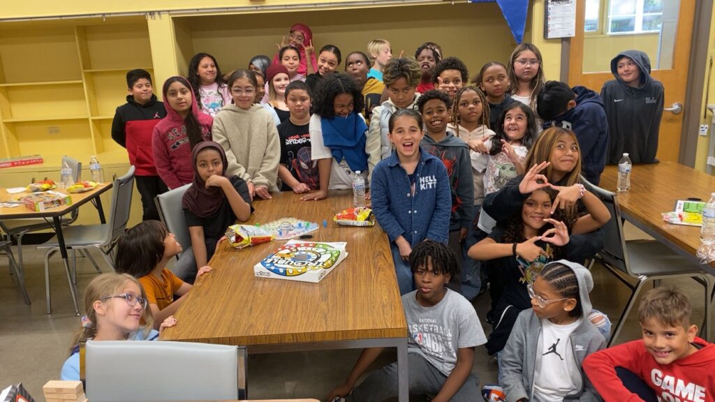 A group of about 30 students stand and sit around a table in a classroom.