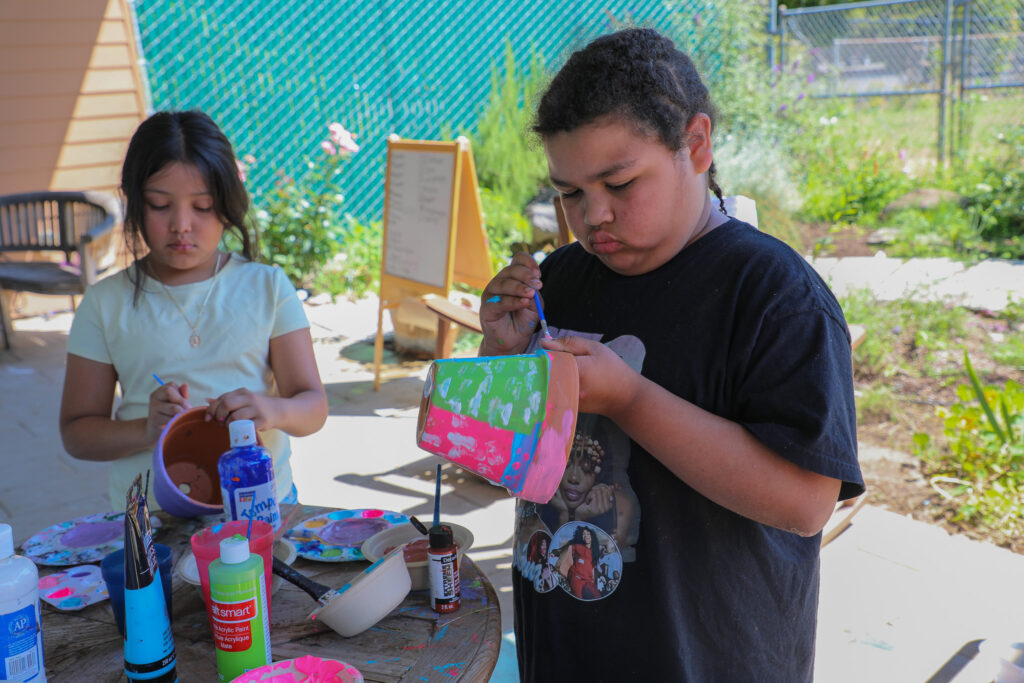 Photo of two children standing around a table covered in paint bottles and art supplies. Both children hold paintbrushes in their right hands to paint flowerpots held in their left hands.