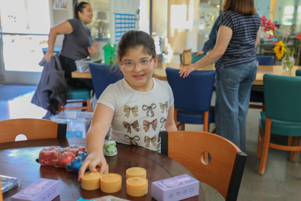 Child wearing white t-shirt and glasses stands in front of a table. Right hand reaches out to choose a squishy toy. Two adults stand at another table in the background.