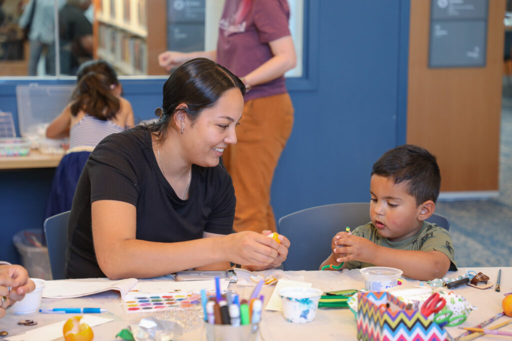 Photo of woman seated on the left in a black t-shirt peeling a mandarin orange. She smiles while looking at her son, who is sitting to her left wearing a green t-shirt while stringing a bead onto his pipe cleaner.