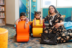 Photo of Maryuri sitting on the floor on the right with their two younger children on the left, who are crouching behind toys.