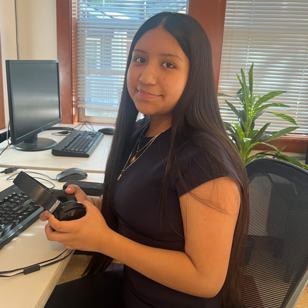 Photo of a student in a black top, holding headphones, and sitting in an office chair in front of a computer keyboard. The student is looking at the camera and smiling.